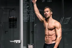 Robert Gryn bez koszulki wskazuje ręką tablicę z wynikami treningów na ergometrze wioślarskim