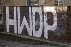Wielki biały napis HWDP namalowany na ceglanym murze przy chodniku, wokół mniejsze niebieskie tagi graffiti