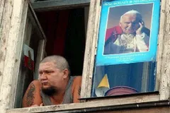 Mężczyzna z tatuażami wygląda przez okno starej kamienicy, obok w szybie wisi plakat z Janem Pawłem II i napisem „Bóg bogaty w miłosierdzie”