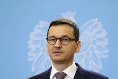 Mateusz Morawiecki w okularach, garniturze i krawacie na tle niebieskiej ścianki z białym orłem