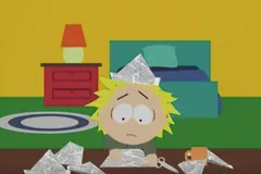 Zestresowany Tweek z serialu South Park siedzi przy biurku wśród papierowych łódek, z gazetową czapką na głowie