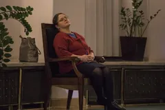 Krystyna Pawłowicz w czerwonym swetrze siedzi na drewnianym krześle z odchyloną głową i zamkniętymi oczami
